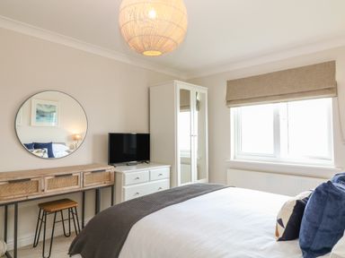 A bedroom with a bed a dresser with a television a wooden desk with a stool and a round mirror at Breakers in Newquay