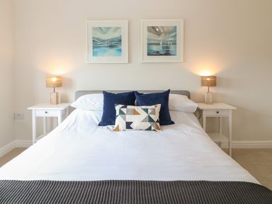 A bedroom with a bed a gray throw pillows two white bedside tables with lamps and two paintings on the wall at Breakers in Newquay