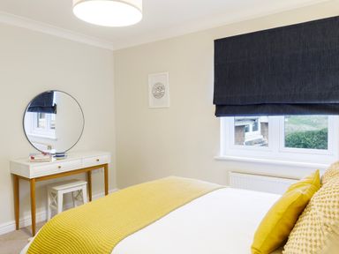 A bedroom with a bed covered by a yellow blanket and pillows next to a window with blue curtains and a vanity table with a round mirror at Breakers in Newquay