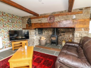 A living room with a stone fireplace wood stove leather sofa wooden table with wine glasses and a lamp on floral wallpaper at Cwmfadog in Llanafan-fawr near Beulah