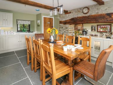 A kitchen with a wooden dining table and chairs a cake and sunflowers on the table a stone wall with a clock and coffee maker at Cwmfadog in Llanafan-fawr near Beulah