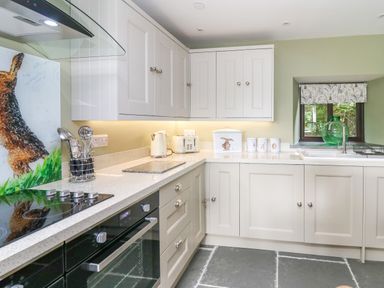 A kitchen with white cabinets an oven a stovetop a kettle a toaster a window with a patterned blind and a green glass jug on the windowsill at Cwmfadog in Llanafan-fawr near Beulah