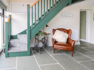 A staircase with green railing and grey carpet next to a brown leather armchair with a white cushion and an antique sewing machine table with a lamp and plant beneath the stairs at Cwmfadog in Llanafan-fawr near Beulah