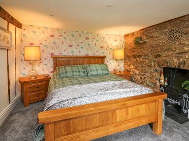 A bedroom with a wooden bed frame green plaid bedding two bedside tables lamps floral wallpaper and a stone wall with a fireplace at Cwmfadog in Llanafan-fawr near Beulah