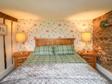 A bedroom with a wooden bed frame green plaid bedding two wooden nightstands with lamps floral wallpaper and a stone wall at Cwmfadog in Llanafan-fawr near Beulah