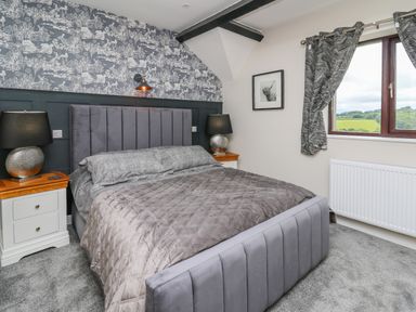A bedroom with a grey upholstered bed grey bedding two bedside tables with lamps a patterned wallpapered wall and a window with curtains at Cwmfadog in Llanafan-fawr near Beulah