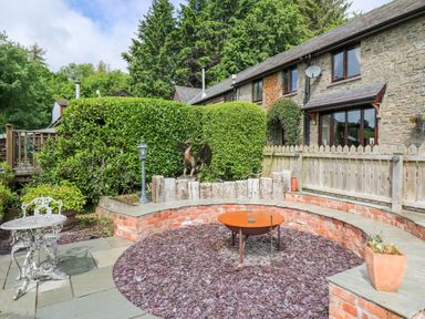 An outdoor patio area with a curved brick bench around a fire pit and a metal table and chairs near a stone house with wooden fence and greenery at Cwmfadog in Llanafan-fawr near Beulah