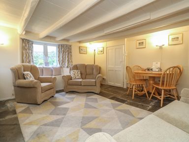 A living room with beige armchairs and sofa a wooden dining table with chairs and a geometric patterned rug at 2 Rose Cottages in Lansallos near Polperro