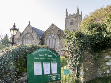 A stone church building with a tower behind a gate and a green notice board at 3 Rose Cottages Lansallos near Polperro