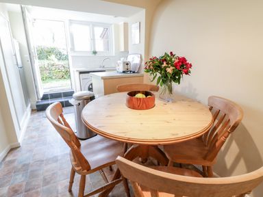 A kitchen with a round wooden table and four wooden chairs a fruit bowl and flowers on the table a door leading outside 3 Rose Cottages Lansallos near Polperro