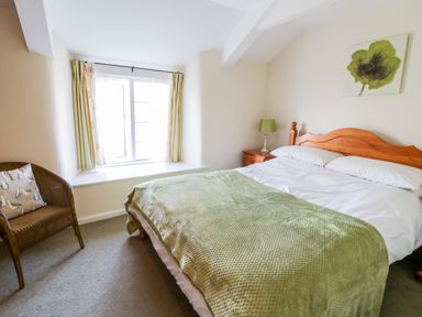 A bedroom with a wooden bed with white linens and green blanket next to a window and a chair at 3 Rose Cottages Lansallos near Polperro
