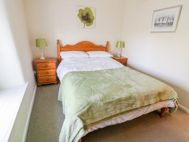 A bedroom with a wooden bed, green blanket, two wooden bedside tables with green lamps and artwork on the walls at 3 Rose Cottages Lansallos near Polperro