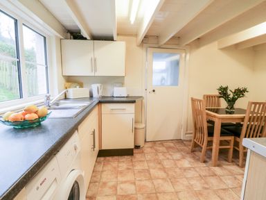 A kitchen with a countertop, sink, fruit bowl, washing machine, white cabinets, small dining table with four wooden chairs and a door at 4 Rose Cottages Lansallos near Polperro