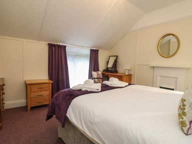 A bedroom with a bed with white sheets and purple throw blankets a dresser a vanity mirror a chair with cushions purple curtains and a round mirror above a fireplace at 4 Rose Cottages in Lansallos near Polperro