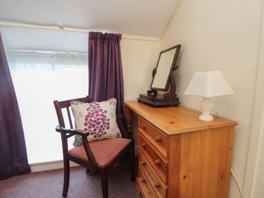 A wooden chair with a cushion next to a wooden dresser with a mirror and lamp near a window with purple curtains at 4 Rose Cottages in Lansallos near Polperro