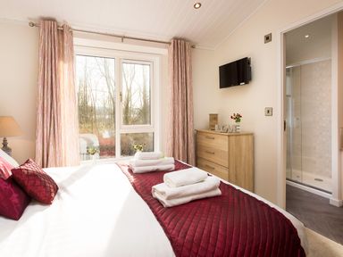 A bedroom with a bed covered in burgundy quilted bedspread and white towels near curtains and window at Waternook Lodge in South Lakeland Leisure Village