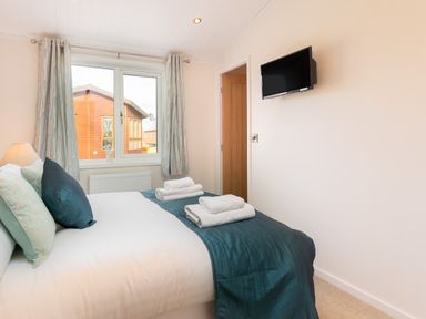 A bedroom with a bed covered with teal bedding and white towels folded on top a window with curtains and a wall mounted tv at Waternook Lodge in South Lakeland Leisure Village