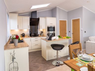 A kitchen with white cabinets a wooden countertop two black bar stools and a dining table with glassware at Waternook Lodge in South Lakeland Leisure Village