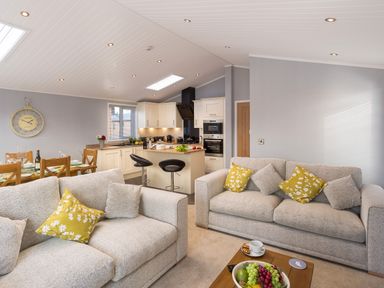 An open living room and kitchen area with beige sofas and yellow cushions at Waternook Lodge South Lakeland Leisure Village