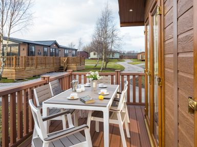 An outdoor deck with a table and chairs set for tea and a book at Waternook Lodge South Lakeland Leisure Village