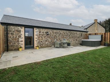An outdoor patio with stone paving a stone wall building a hot tub two chairs and a table on the patio and grass in the foreground at The Cwtch in St Brides Major