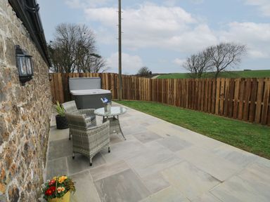 An outdoor patio with two wicker chairs a glass table a hot tub a wooden fence and grass at The Cwtch in St Brides Major