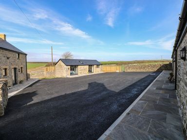 A paved driveway between stone buildings with fields in the background at The Cwtch in St Brides Major