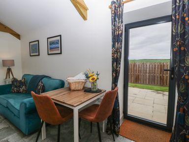 A small dining area with a wooden table and two chairs next to a blue sofa and a glass door with floral curtains at The Cwtch in St Brides Major