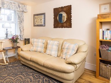 A living room with a beige leather sofa with three checked cushions a wooden bookshelf a small wooden table with flowers and lamp and a window with patterned curtains at Old Post Office in Bull Bay