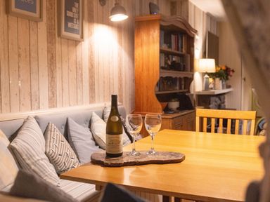 A dining area with a wooden table a wine bottle and two glasses on a wooden tray a cushioned bench with pillows and a wooden bookshelf in the background at Old Post Office in Bull Bay