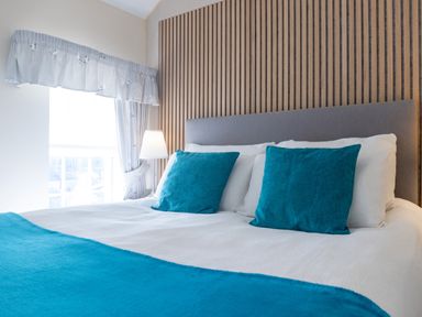 A bed with blue pillows and a matching blanket beside a window with curtains and a lamp at Old Post Office in Bull Bay