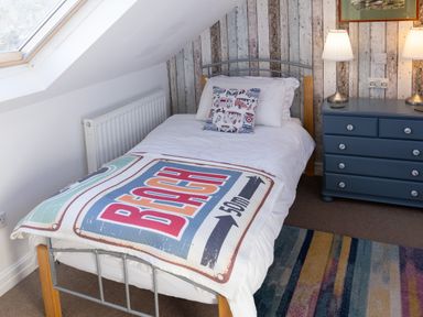 A bedroom with a single bed with a blanket featuring the word beach a bedside chest with two lamps and a striped rug at Old Post Office in Bull Bay