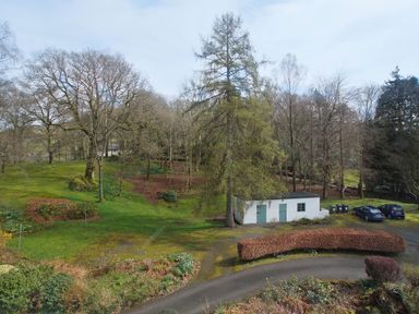 A green lawn with trees a curved hedge a small white building and parked cars at Sethera in Ambleside