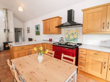 A kitchen with wooden cabinets a red stove a wooden dining table with flowers a wood burning stove and two paintings on the wall at Y Buarth in Waunfawr