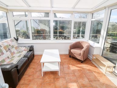 A sunroom with a brown sofa covered with a quilt a brown armchair a white coffee table and a wooden shoe rack at Y Buarth in Waunfawr