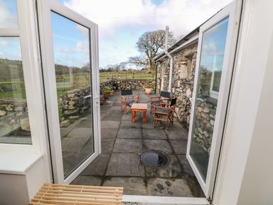 An outdoor patio with stone flooring wooden chairs and a table with stone walls and a field in the background at Y Buarth in Waunfawr