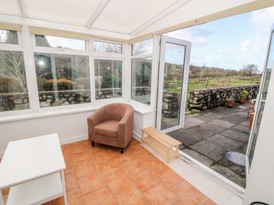 A conservatory with a brown armchair a white table and a wooden shoe rack at Y Buarth in Waunfawr