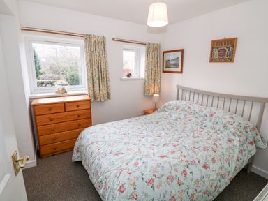 A bedroom with a bed with floral bedding a wooden chest of drawers and two windows at Y Buarth in Waunfawr