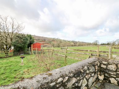 A grassy garden area with a bird bath a small tree a stone wall a red shed and a view of fields and hills at Y Buarth in Waunfawr