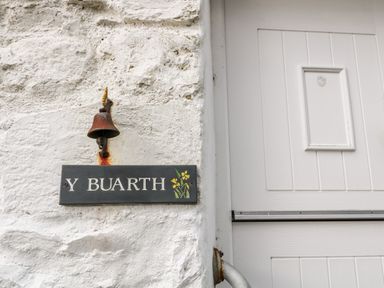 A white textured wall with a rusted metal bell and a sign reading Y Buarth next to a white paneled door at Y Buarth in Waunfawr