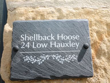 A black rectangular plaque with white text mounted on a textured stone wall reading Shellback Hoose 24 Low Hauxley at 24 Hauxley Links in Low Hauxley near Amble