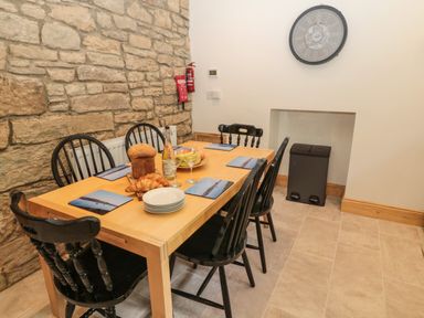 A dining area with a wooden table set with plates croissants and bread six black chairs stone wall and clock on a white wall at 24 Hauxley Links Low Hauxley near Amble