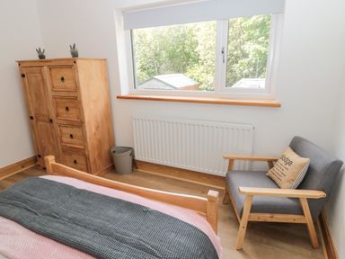 A bedroom with a wooden bed, wooden wardrobe, armchair with cushion, and a window overlooking trees at 24 Hauxley Links in Low Hauxley near Amble