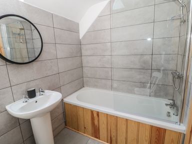 A bathroom with a pedestal sink a round mirror a bathtub with glass shower screen and gray tiled walls at 24 Hauxley Links in Low Hauxley near Amble