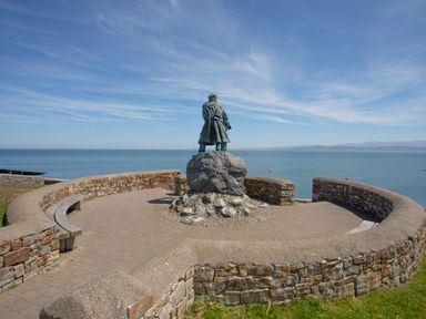 A statue on a stone pedestal enclosed by a curved stone wall overlooking the sea at Bryn Peris in Moelfre