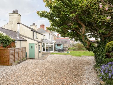 A garden with gravel driveway a tree green lawn outdoor table and chairs and houses in the background at Bryn Peris in Moelfre