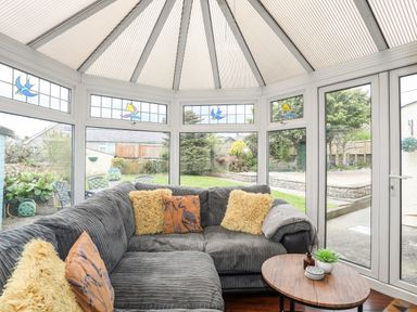 A conservatory with a grey corner sofa with assorted cushions and a small wooden table with decorative items at Bryn Peris in Moelfre