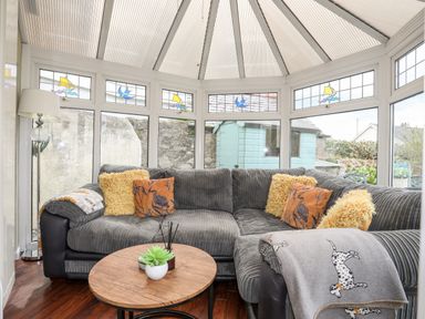 A conservatory with a grey sofa yellow and orange pillows round wooden table and a standing lamp at Bryn Peris in Moelfre