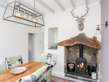 A dining area with a wooden table and floral chairs next to a stone fireplace with a wood stove and a metallic deer head on the wall at Bryn Peris in Moelfre