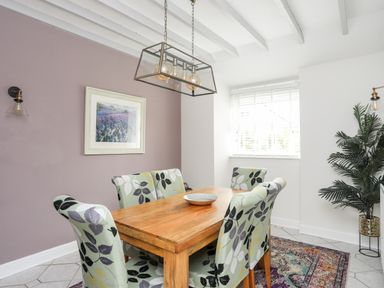 A dining room with a wooden table six floral upholstered chairs a framed picture on a purple wall a hanging light fixture and a large plant at Bryn Peris in Moelfre